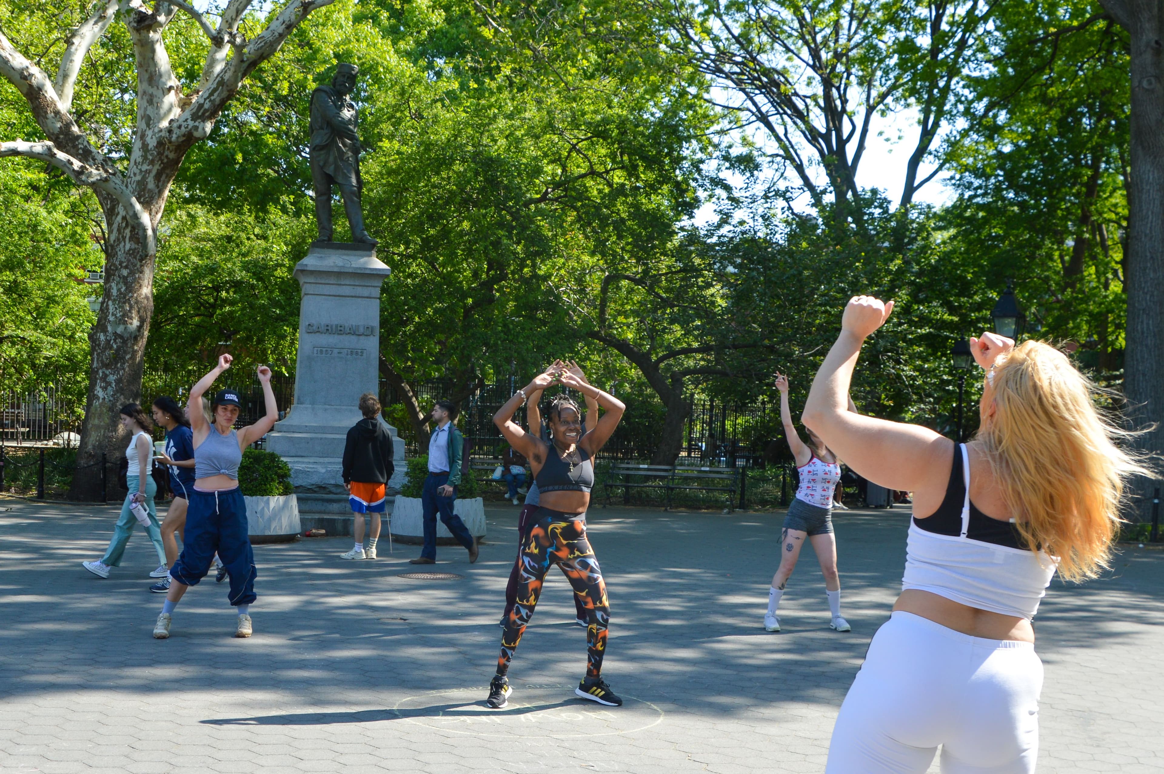 People follow an instructor and dance at Bodyroll in Washington Square Park with a statue of Garibaldi in the background.