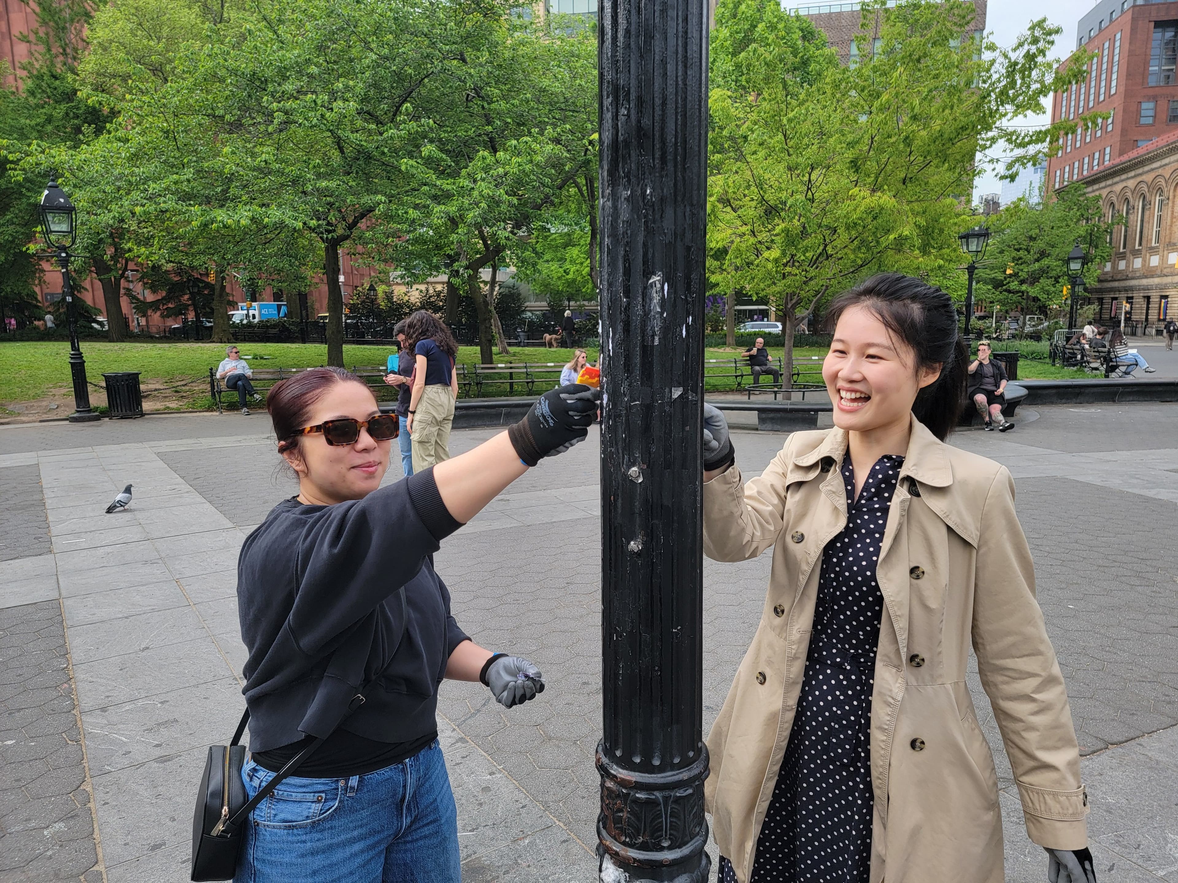 Two people scrape stickers off of a lamppost in Washington Square Park.