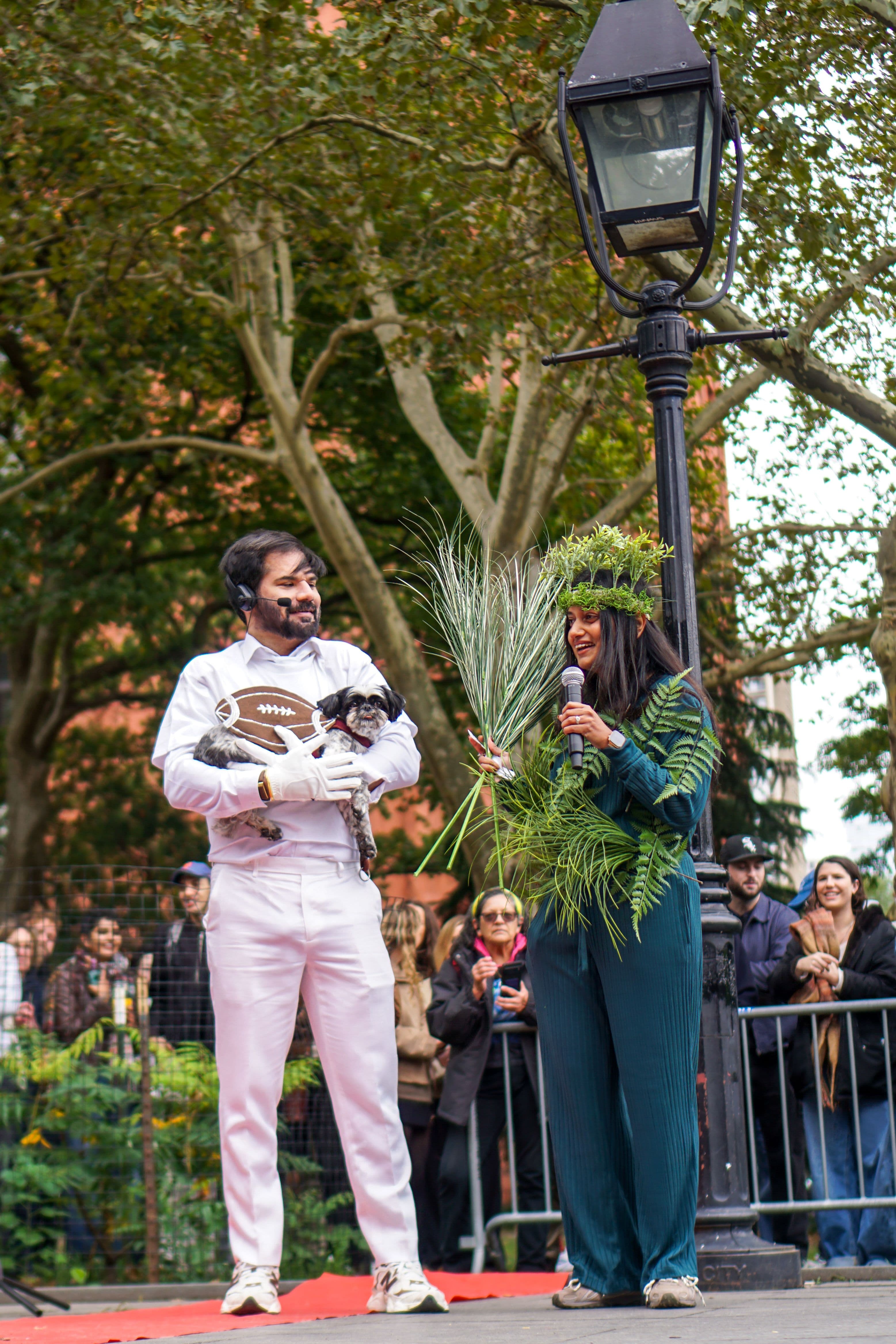 A man dressed all in white with a headset microphone holds a small black-and-white dog wearing a football costume on a red carpet in Washington Square Park. Beside him, a woman wearing ferns and a leafy crown holds a microphone and a bundle of tall grasses as spectators watch from behind barricades.