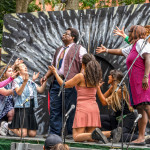 a group of performers from Theater for the New City on stage in the park. The stage has microphones and a painted backdrop.
