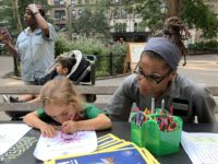 the Youth Activities Coordinator, Miss Debbie, watches a child use crayons on a coloring page