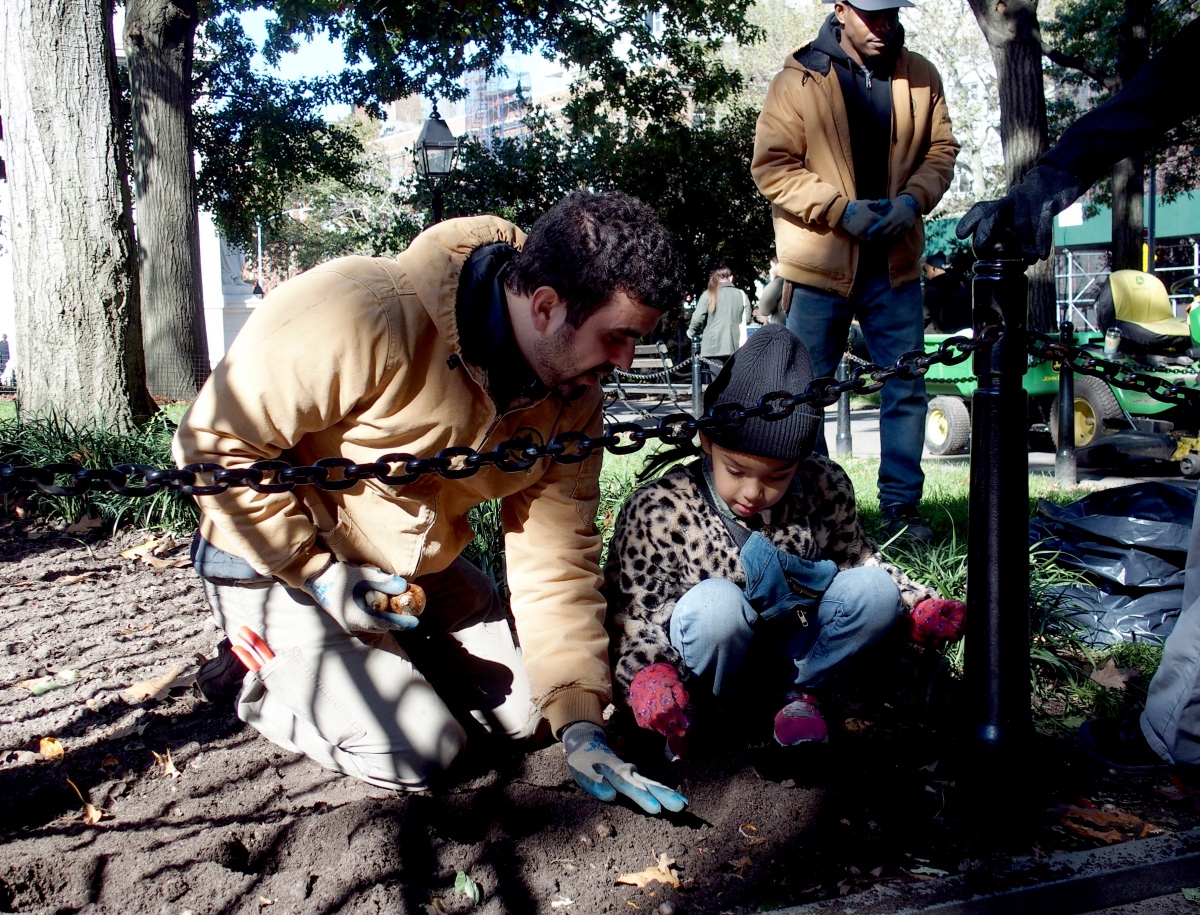 a gardener kneels in the dirt and demonstrates a gardening technique to a child