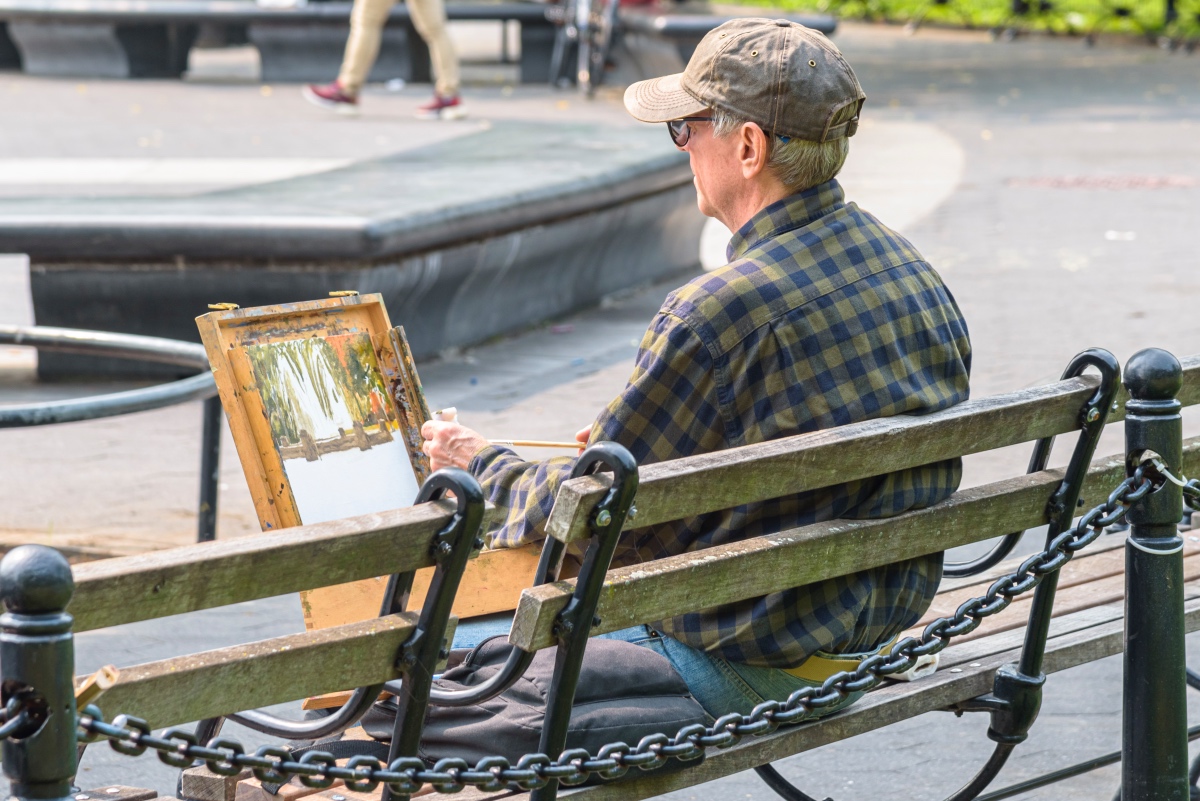 man wearing a plaid shirt and a baseball hat sits on a bench with his back to the camera while painting a picture of the fountain