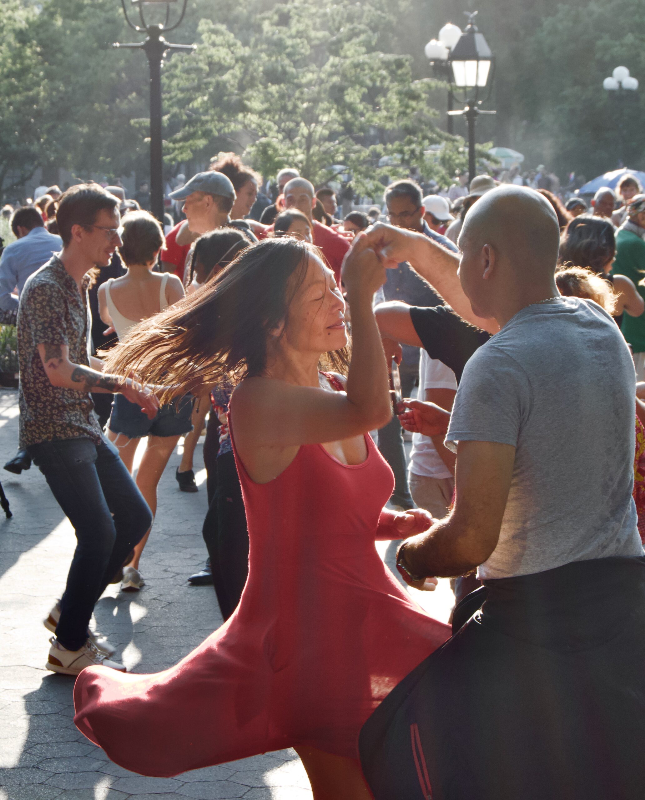 a crowd of people dancing in the park featuring a woman wearing a red dress being spun by a dance partner