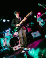 blurry photo of three band members performing on stage, center person in focus stands with an electric guitar