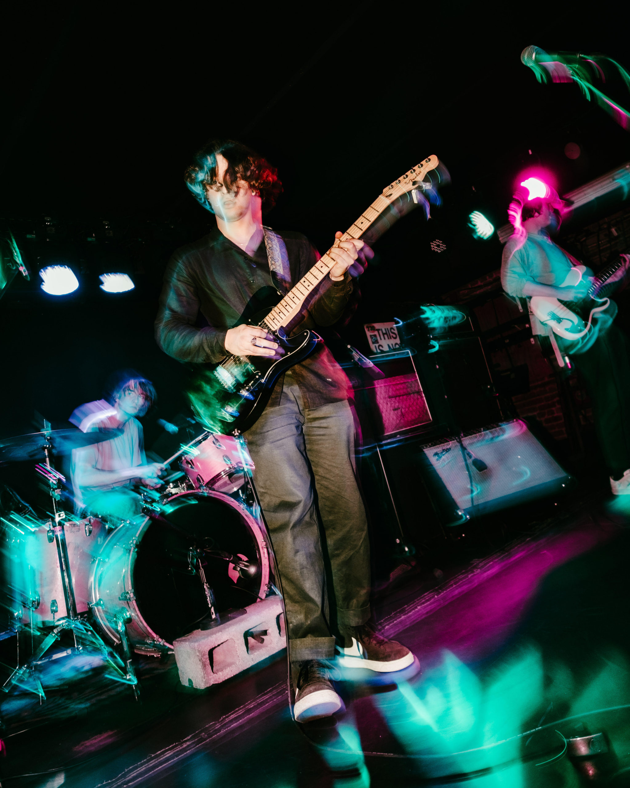 blurry photo of three band members performing on stage, center person in focus stands with an electric guitar