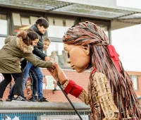 three children hold Little Amal's hand in the park