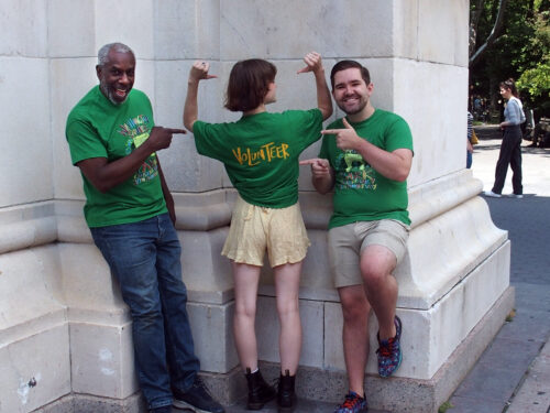 three volunteers wear green washington square park shirts, one person in the middle has back turned to camera showing the word volunteer on the back of shirt, all three volunteers are pointing to the back of the shirt