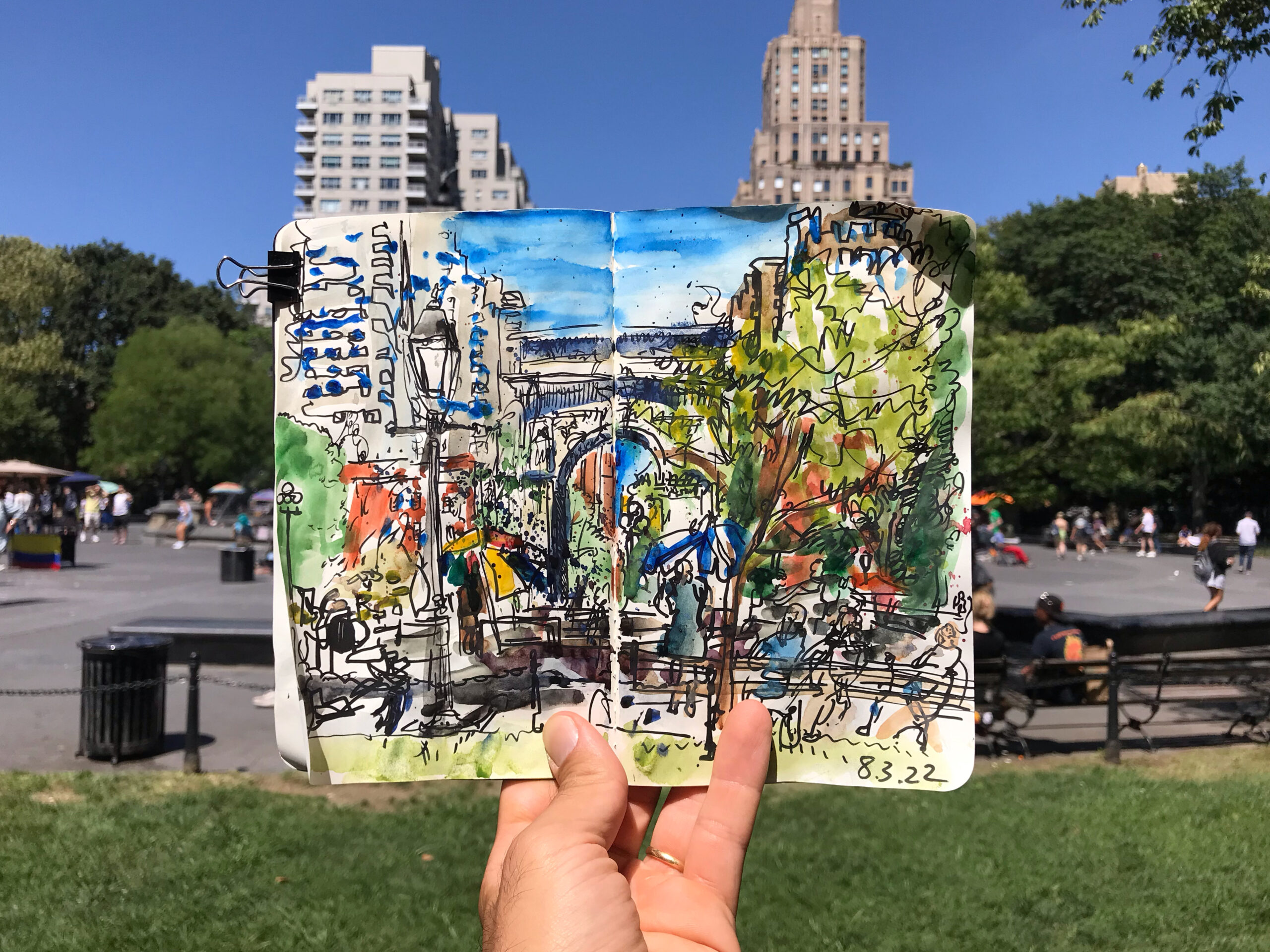 hand holding a sketchbook open to a watercolor image of the arch, trees, and people sitting on park benches