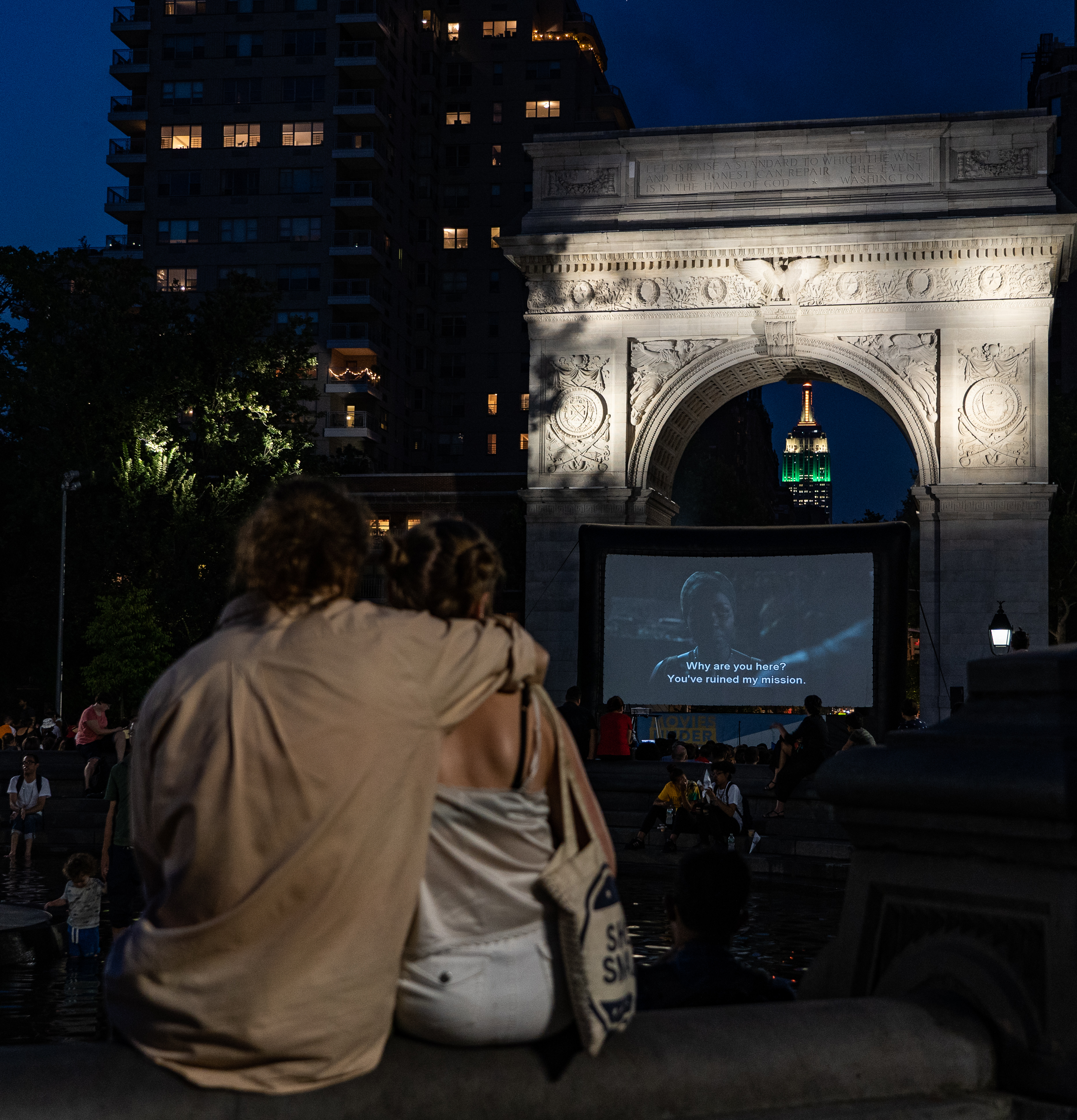 a couple sits together on the edge of the Washington Square Park Fountain watching a french film on a large screen under the Arch